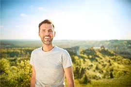 Man in grey shirt smiling while outside with green background