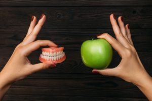 Hands at wood table holding a set of dentures and a green apple